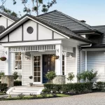 Front view of light gray Tudor-style house with dark tile roof, white trim, porch, and manicured garden